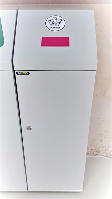 GEMOS, a specialist in waste separation systems, has developed a waste bin for discarded mobile phones on behalf of the climate protection management. The picture shows the waste bin. A tall, light grey container with a flap.